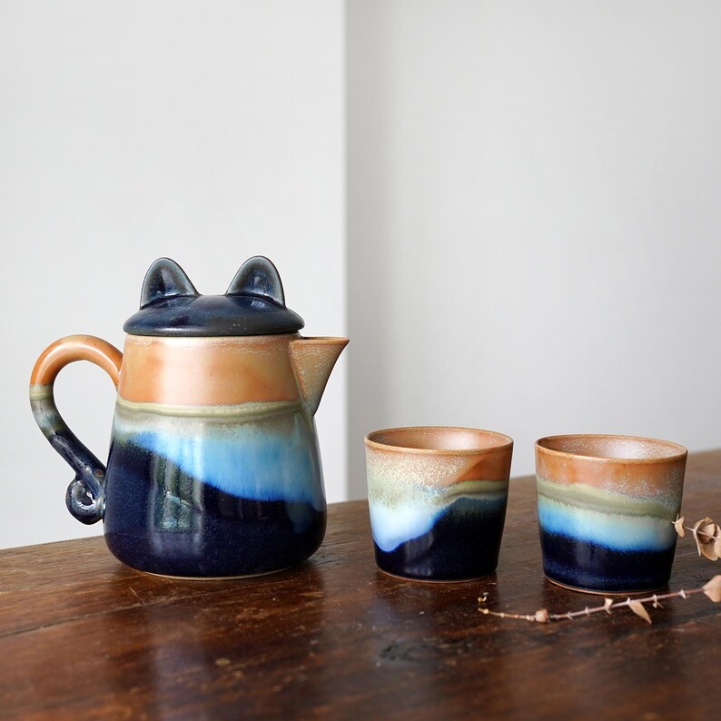 May include: A ceramic teapot with a cat-ear lid, a handle, and a spout, alongside two matching cups. The teapot and cups feature a color gradient of blue, orange, and brown. The set is on a wooden surface.