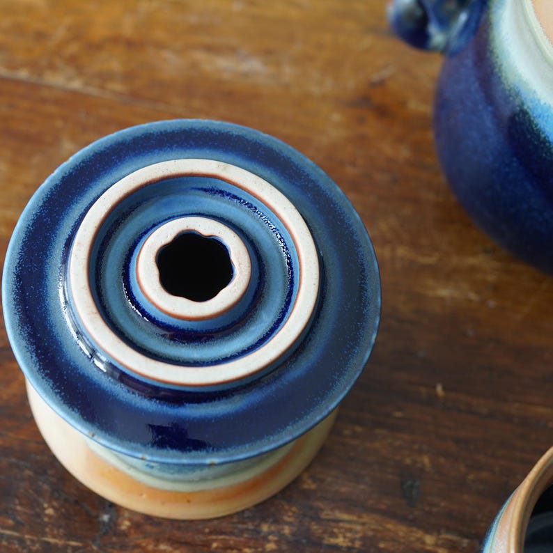 May include: A ceramic butter dish with a deep blue lid and a cream-colored interior. The lid has concentric circles and a central opening. The base has a gradient of colors, including beige, orange, and blue. The dish is on a wooden surface.