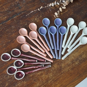 May include: A selection of ceramic spoons in shades of pink, purple, and white, displayed on a wooden surface. Some spoons feature a dark red filling. The spoons vary in size and shape, ideal for serving or tasting.