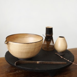 Könnte beinhalten: Eine beige Matcha-Schale aus Keramik mit Ausguss, ein Bambusbesen, eine kleine Vase und ein Bambuslöffel, alles auf einem dunklen Holztablett angeordnet. Für die Zubereitung von Matcha-Tee. Die Schale hat einen dunklen Rand.