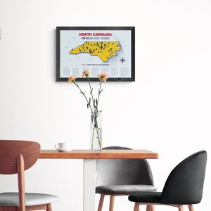 May include: A framed map of North Carolina with the text "North Carolina Top 50 Disc Golf Courses" printed on it. The map is yellow and shows the location of 50 disc golf courses in the state. The map is hanging on a white wall above a wooden table with three chairs.