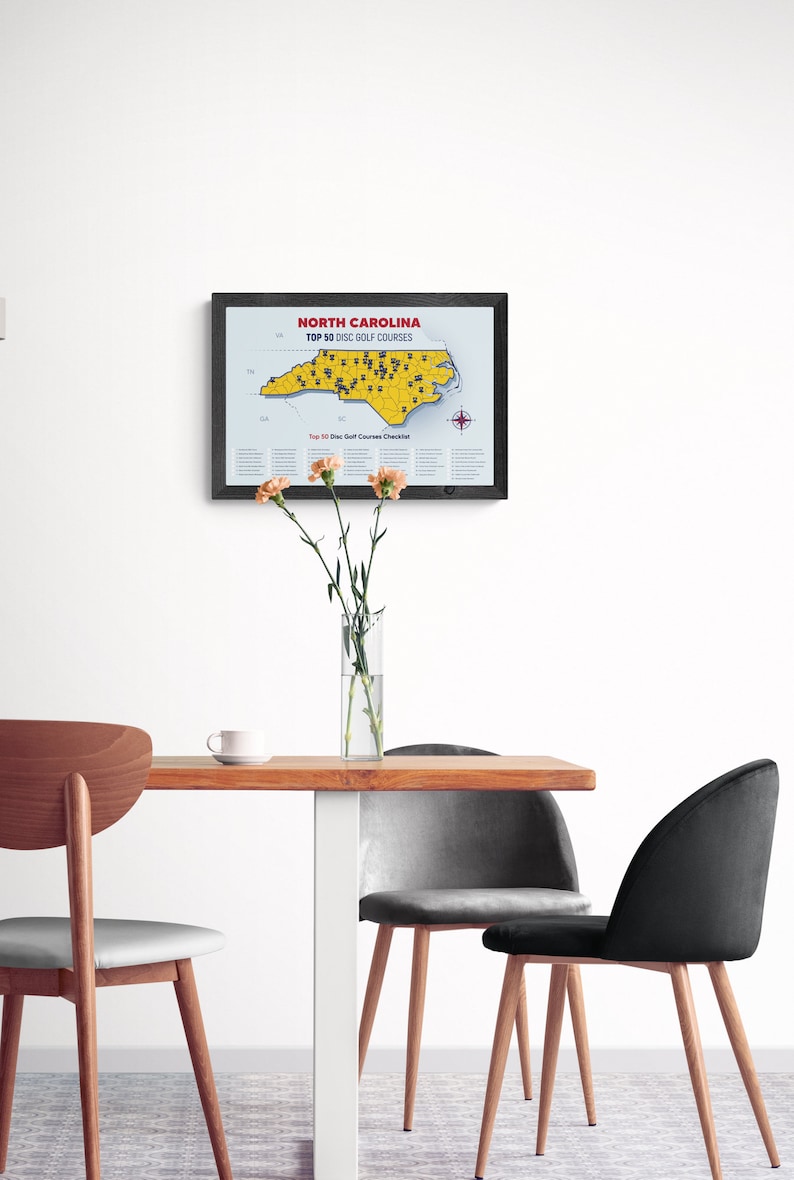 May include: A framed map of North Carolina with the text "North Carolina Top 50 Disc Golf Courses" printed on it. The map is yellow and shows the location of 50 disc golf courses in the state. The map is hanging on a white wall above a wooden table with three chairs.