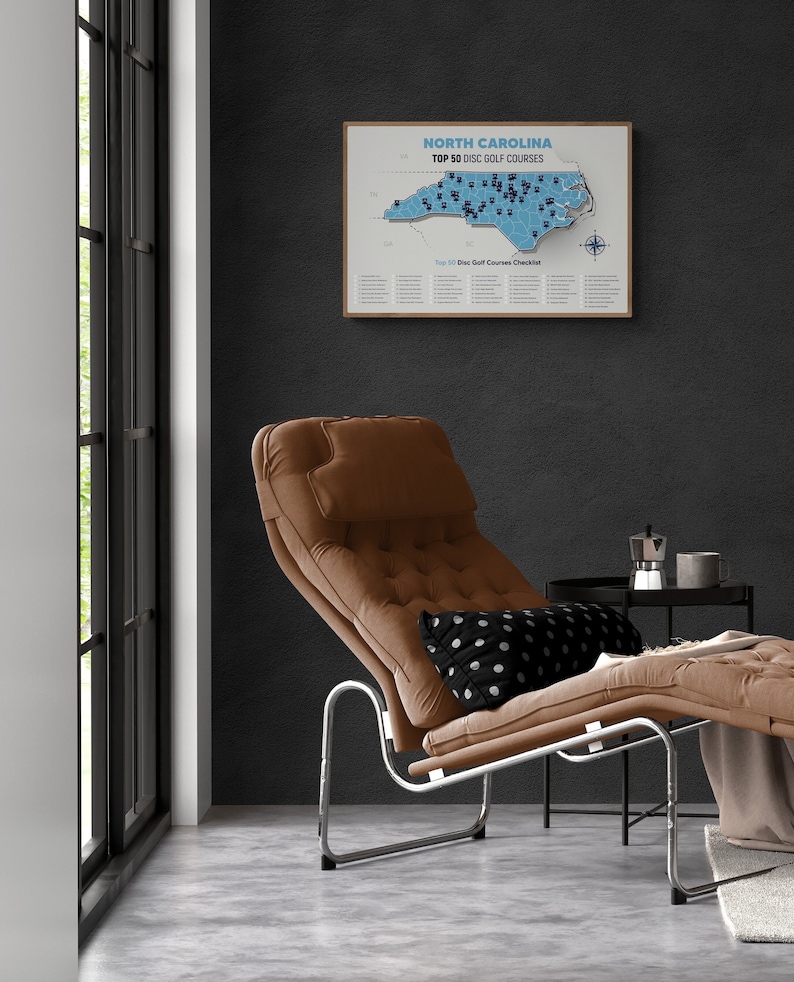 May include: A framed map of North Carolina with the top 50 disc golf courses marked. The map is hanging on a dark gray wall above a brown leather chaise lounge with a black and white polka dot pillow.