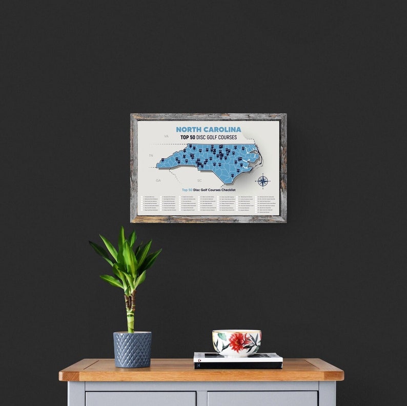 May include: A framed map of North Carolina with a blue outline of the state and black dots marking the locations of the top 50 disc golf courses. The title of the map reads "North Carolina Top 50 Disc Golf Courses".