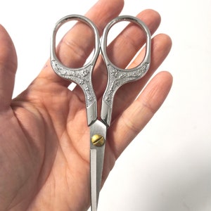 Könnte beinhalten: Silberfarbene Dekorationsschere mit floralem Muster auf den Griffen. Die Schere hat eine goldfarbene Schraube in der Mitte.