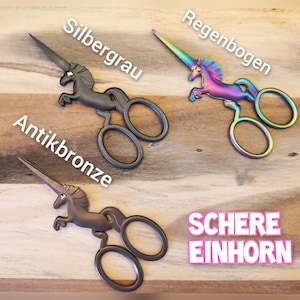 Könnte beinhalten: Drei unicornförmige Scheren mit verschiedenen Farbtönen: Antikbronze, Silbergrau und Regenbogen. Die Scheren liegen auf einer Holzoberfläche mit dem Text "SCHERE EINHORN" darunter. Der Text über jeder Schere lautet "Antikbronze", "Silbergrau" und "Regenbogen".