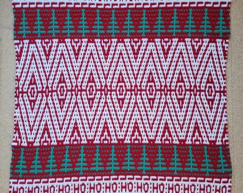 May include: A red, white, and green crocheted blanket with a Christmas-themed design. The blanket features a repeating pattern of trees, diamonds, and the words "HO HO HO".