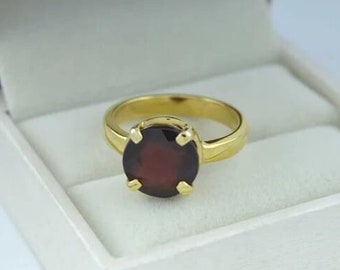 Puede incluir: Un anillo de oro con una piedra ovalada de color rojo oscuro engastada en garras. El anillo se muestra en una caja de anillo blanca.