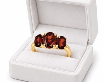 May include: A gold ring featuring three oval-cut deep red garnet gemstones set in a prong setting. The center stone is larger than the two flanking stones. The ring is displayed inside an open, white, velvet-lined jewelry box with a hinged lid.