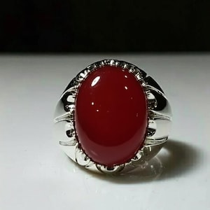 May include: A silver ring featuring a large, oval, deep red gemstone. The ring's band has a decorative design around the gemstone. The polished gemstone reflects light, giving it a glossy appearance. The ring is set against a white surface.