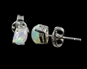May include: A pair of stud earrings with oval-shaped opals set in silver. The opals display a play of color with iridescent hues of green, blue, and yellow. The earrings are set against a black background.