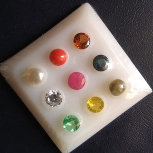 May include: A white tray with nine gemstones, including a red coral, a white pearl, a ruby, a diamond, an emerald, a yellow sapphire, a blue sapphire, a green peridot, and a brown topaz.