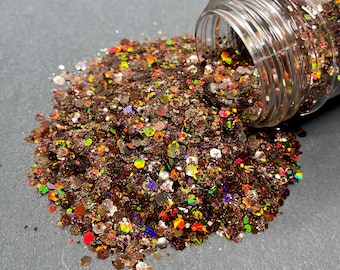 Op de afbeelding: Een glazen pot met een bruine, iriserende glittermix die op een grijs oppervlak wordt gegoten. De glitter is een mix van bruin, goud, oranje, rood, groen en blauw.