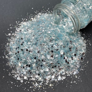 May include: A close-up of a jar of light blue and silver glitter. The glitter is spilling out of the jar onto a black surface.