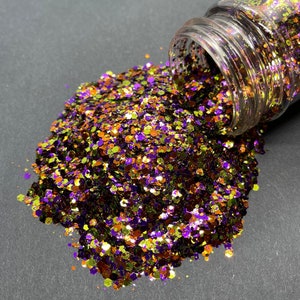 May include: A clear jar filled with a mixture of purple, gold, orange, and green glitter. The glitter is spilling out of the jar onto a dark gray surface.