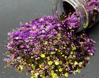 Op de afbeelding: Een glazen pot gevuld met paarse en gouden glitter, die uitloopt op een grijs oppervlak.