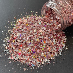May include: A clear jar tipped over, spilling a mix of star-shaped glitter in shades of pink, silver, and iridescent colors. The glitter is scattered on a dark surface, creating a sparkling effect. Ideal for crafts and decorations.