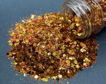 Puede incluir: Un tarro de purpurina dorada y roja se derrama sobre una superficie gris oscuro. La purpurina es una mezcla de piezas pequeñas y grandes, creando un aspecto brillante y festivo.