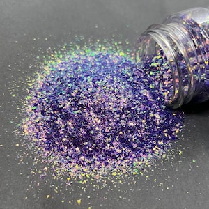 May include: A clear glass jar filled with iridescent purple and green glitter. The glitter is spilling out of the jar onto a dark gray surface.