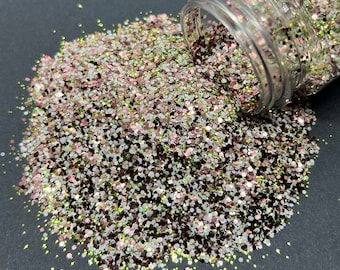Op de afbeelding: Een glazen pot gevuld met een mengsel van groene, roze, witte en gouden glitter. De glitter stroomt uit de pot op een zwart oppervlak.