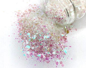 May include: A clear glass jar tipped over, spilling iridescent glitter onto a white surface. The glitter is a mix of white, pink, and teal, creating a sparkling effect. The jar's lid is visible, and the glitter is scattered around it.
