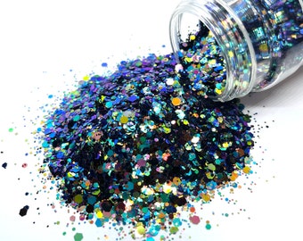 May include: A clear glass jar filled with iridescent blue, green, gold, and black glitter. The glitter is spilling out of the jar onto a white surface.