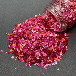 May include: A clear jar tipped over, spilling a vibrant mix of pink, red, and orange confetti-like glitter. The glitter is in various sizes and shapes, creating a textured, sparkling effect. The background is a dark gray surface.