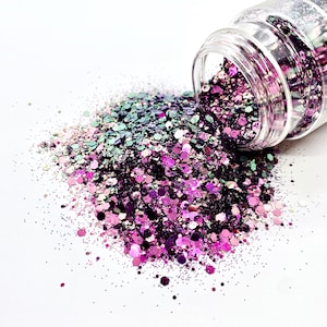 May include: A clear glass jar filled with iridescent glitter in shades of green, purple, and pink. The glitter is spilling out of the jar onto a white surface.