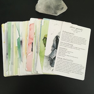 May include: A collection of oracle cards with watercolor illustrations and text, alongside a clear quartz crystal. The cards feature various colors and imagery, with one card displaying the text "clear quartz". The cards are on a dark surface.