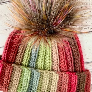 May include: A crocheted beanie with a multi-coloured striped pattern in shades of red, green, and brown. The beanie has a fluffy pom pom on top with brown, pink, and yellow streaks.