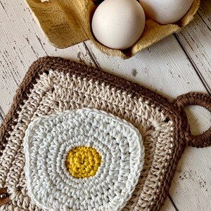 May include: A crochet potholder featuring a fried egg design. The square potholder has a brown border and a beige centre. The fried egg is white with a yellow yolk. Three white eggs are in a cardboard egg box.