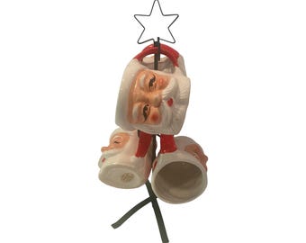 Puede incluir: Un juego de tres tazas de cerámica de Papá Noel, cada una con una cara blanca, una bufanda roja y un gorro blanco. Las tazas están suspendidas de un soporte de metal negro rematado con una estrella. Las tazas están diseñadas para las vacaciones de Navidad.