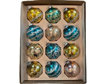 May include: A set of twelve round glass Christmas ornaments arranged in a cardboard box. The ornaments feature metallic finishes in yellow, teal, and pink, decorated with silver glitter patterns including swirls, zigzags, and floral motifs.