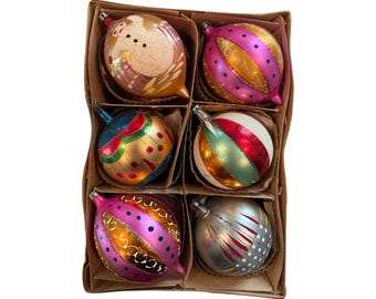 Könnte beinhalten: Ein Set aus sechs Weihnachtskugeln aus Glas in einem Karton. Die Kugeln zeigen verschiedene Designs, darunter einen goldenen Schneemann, rosa und goldene Streifen, eine blaue Kugel mit roten Punkten, eine weiß-rote Kugel und eine silberne Kugel.