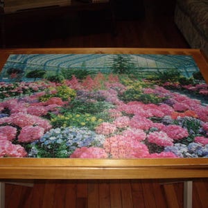 May include: A completed jigsaw puzzle depicting a greenhouse filled with pink and purple flowers. The puzzle is framed in a wooden frame and sits on a wooden table.