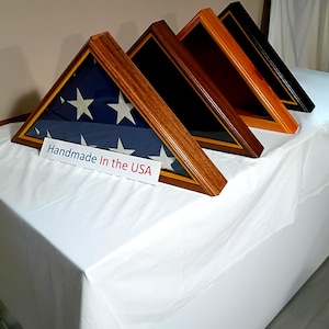 May include: Four triangular wooden display cases arranged in a row. The front case holds a folded American flag. A white rectangular sign in front of the cases features the text "Handmade In the USA" in blue and red lettering on a white background.