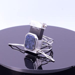 Può includere: Un anello in argento con una pietra rettangolare blu scuro maculata incastonata in una montatura in metallo. La fascia dell'anello è composta da più fili d'argento sottili e sovrapposti. Una chiavetta USB SanDisk nera è integrata nel design.