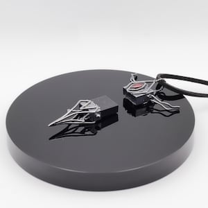 May include: Two silver-toned pendant necklaces with dark accents and red gemstone details rest on a black, circular display. One pendant has a raven skull design, the other a geometric shape. The necklaces are displayed against a white background.