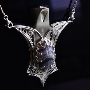 May include: A silver necklace with a detailed filigree design. The pendant features a silver case with the word "SAMSUNG" engraved on it, and a purple gemstone.
