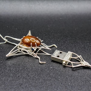 May include: A silver necklace with a star-shaped pendant featuring a brown gemstone and a USB drive. The pendant is designed to resemble a spider web.