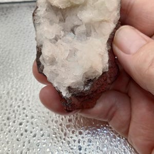 May include: A natural mineral specimen featuring a cluster of white crystals within a reddish-brown matrix. The rough, textured surface of the stone is held in a hand, showcasing its organic form and color variations. This geological item is ideal for collectors.