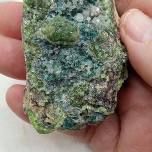May include: A close-up of a mineral specimen with a rough, textured surface. The stone features a mix of green, blue, and white crystalline formations. The mineral is held in a hand, showcasing its natural beauty.