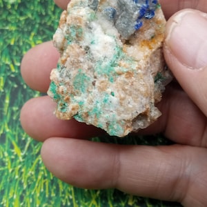 May include: A close-up of a mineral specimen held in a hand. The rock features a mix of colors, including beige, white, green, and blue. The green appears to be malachite, and the blue is likely azurite. The specimen is rough and textured.