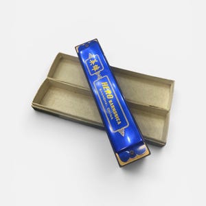 Puede incluir: Una armónica azul con letras doradas que dicen HERO HARMONICA SHANGHAI, CHINA, apoyada en diagonal sobre su caja de almacenamiento rectangular de cartón de color claro. El instrumento presenta un acabado metálico y bordes dorados decorativos.