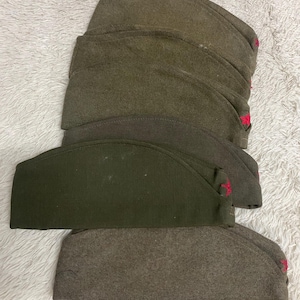 May include: A stack of olive drab military garrison caps with red accents. The caps are made of a wool-like material and are arranged on a white, textured surface. The caps have a classic, vintage design.
