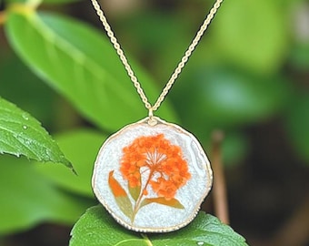 May include: A gold-colored chain necklace with a round pendant. The pendant features a preserved orange flower encased in clear resin with a gold-colored border. The background is blurred green foliage.