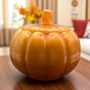 Puede incluir: Un tarro decorativo con forma de calabaza de color naranja con tapa. El tarro tiene un acabado brillante y está sobre una superficie de madera. La calabaza tiene un tallo en la parte superior y está diseñada con surcos verticales.