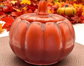 Puede incluir: Un recipiente de cerámica con forma de calabaza con tapa, en tonos naranja y rojo. El recipiente tiene un diseño acanalado y un asa en forma de tallo. El fondo presenta hojas de otoño coloridas y una pequeña calabaza amarilla.