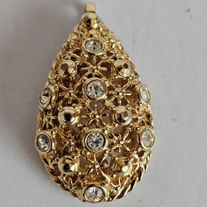 May include: A gold-toned pendant with a teardrop shape. The pendant features a detailed openwork design with small, clear crystals and gold-colored accents. The overall aesthetic is ornate and elegant, suitable for jewelry.