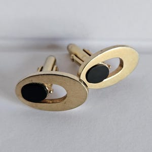 May include: A pair of gold-toned cufflinks with an oval design. Each cufflink features a black, oval-shaped insert. The cufflinks have a classic, elegant appearance, suitable for formal wear.
